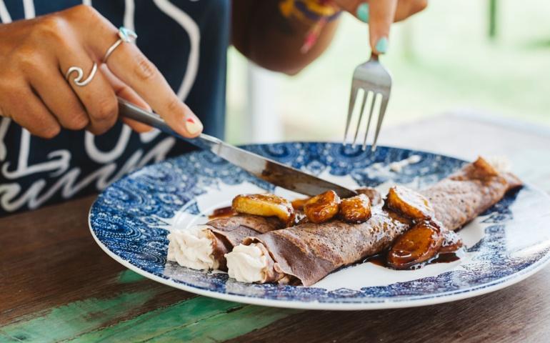 Young woman eating crepes with bananas and creme in outdoor cafe at summer time, France