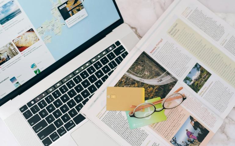 A Top View of Travel Planning Essentials on a Laptop with a Map, Credit Cards, and a Magazine