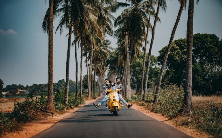A joyful couple rides a yellow scooter through a scenic palm-lined road in Goa