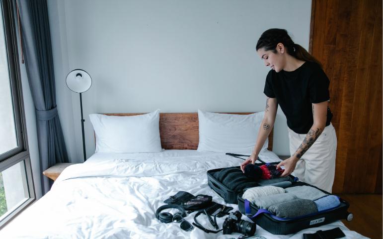 Woman packing suitcase with clothes and camera in a bright bedroom.