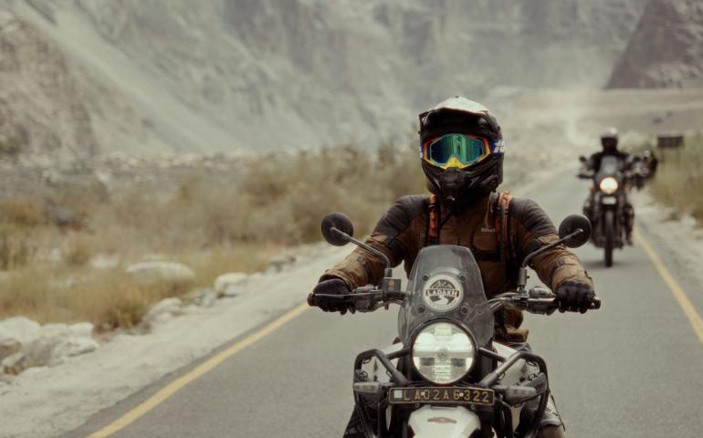 A motorcyclist rides through the scenic Leh mountains under a cloudy sky.