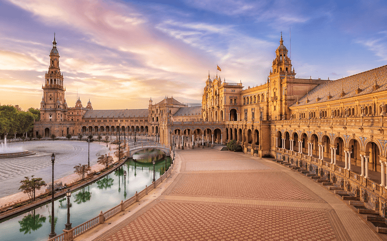 Plaza de España in Seville, Spain, with ornate historic buildings, towers and bridge at sunset.