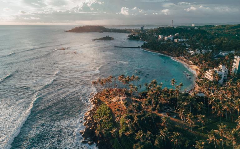 View of Sri Lankan coastline with waves, cliffs, and lush palm trees at sunset