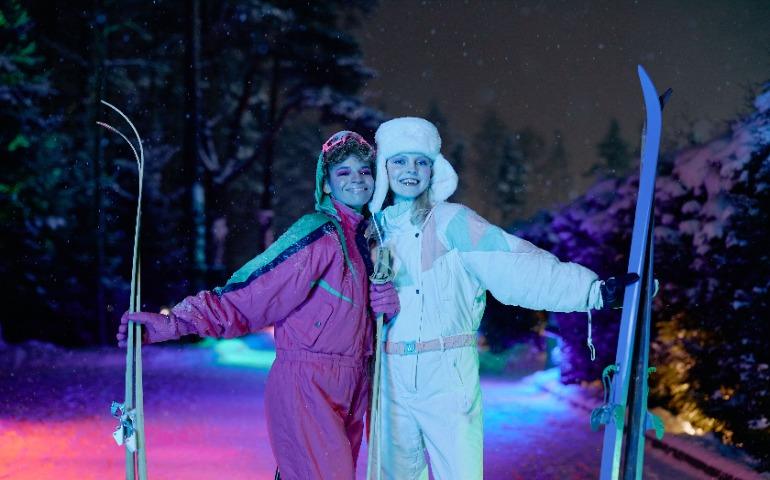 Two women enjoy night skiing under vibrant neon lights, showcasing stylish winter fashion