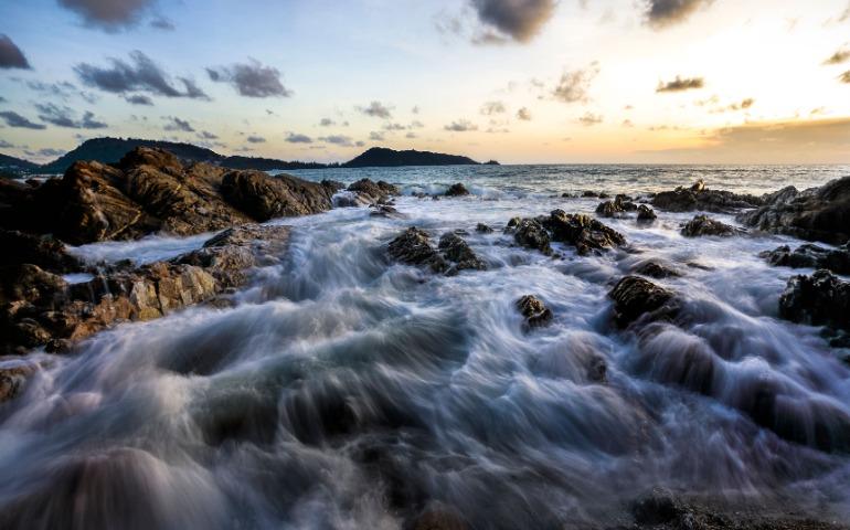 View of waves crashing on a rocky shore in Phuket, Thailand at sunset.